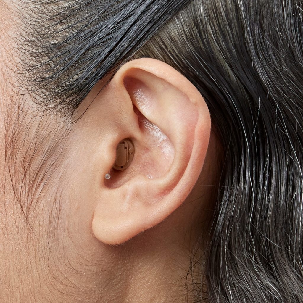Medium-brown Oticon Own SI completely-in-canal hearing aid worn in a woman’s ear